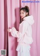 A woman in a pink hoodie leaning against a pink curtain.